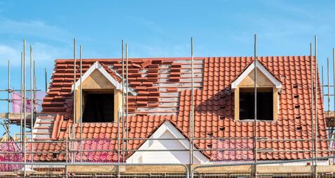 Scaffold poles surrounding the partly tiled roof with dormer windows of a new bungalow being built, against a blue sky.