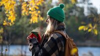 Pleased dreaming woman warms hands on thermos cup outside on cold late autumn, strolling in forest. Nature therapy.